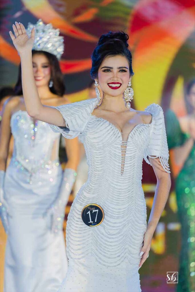 Misty D. Allen smiling and waving to the audience in an embellished white gown with structured sleeves and large earrings