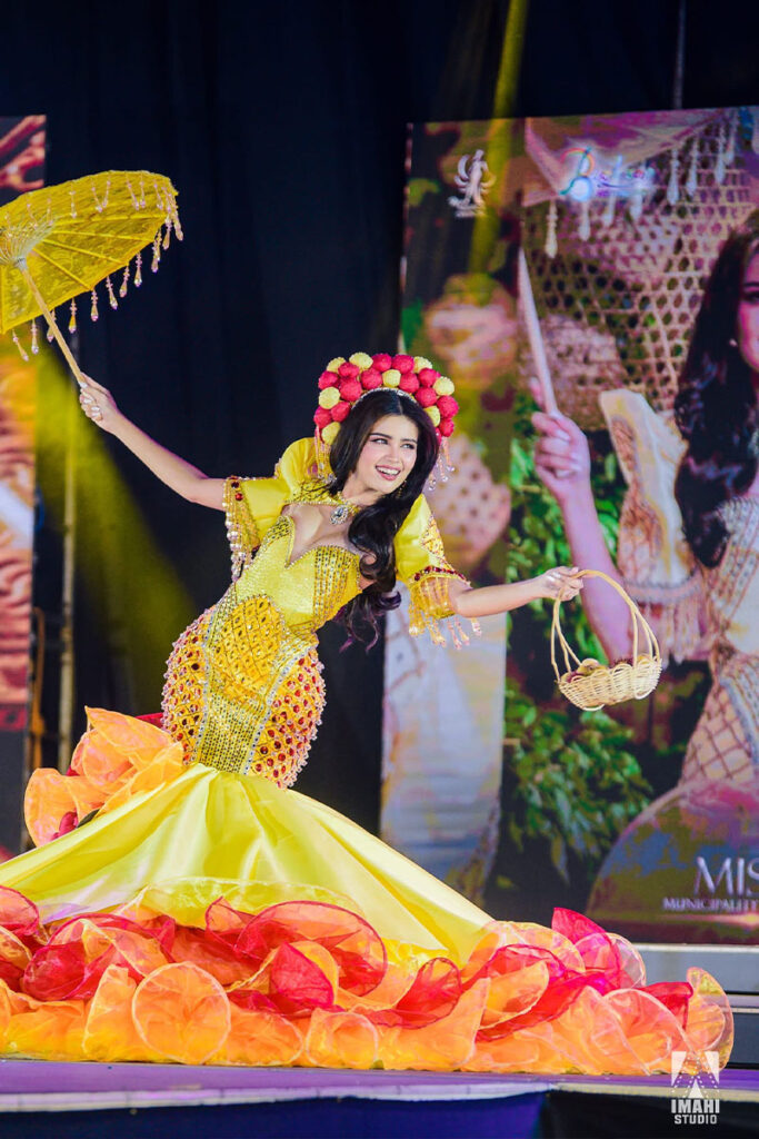 Misty D. Allen performing on stage in a bright yellow festival costume with floral decorations, holding a traditional parasol and basket