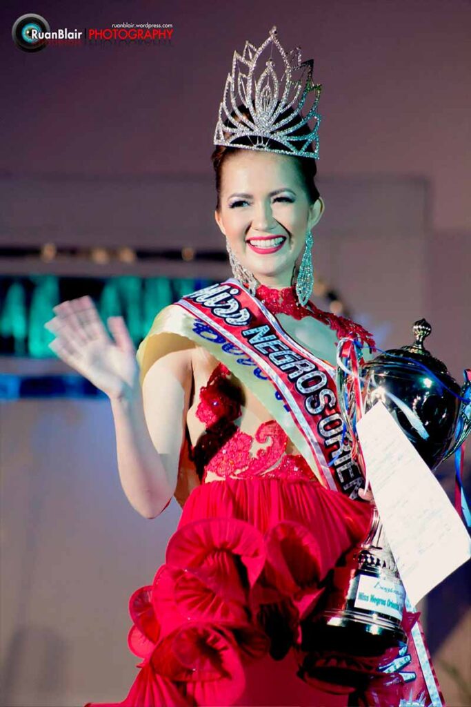 Monica Infante Bartsch waving to the audience in a red evening gown while holding her Miss Negros Oriental Tourism 2013 trophy