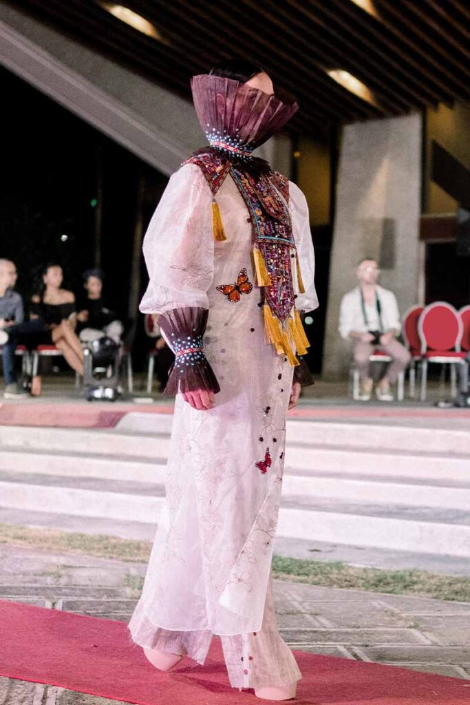 Model in a sheer dress with a high pleated collar, embellished textile details and butterflies from Amir Sali’s collection