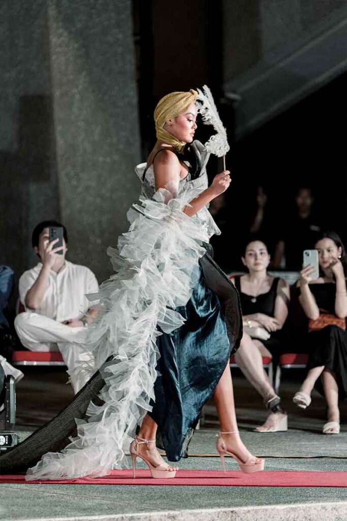 Model in a Tingting Delfin runway look with layered white ruffles and a decorative fan