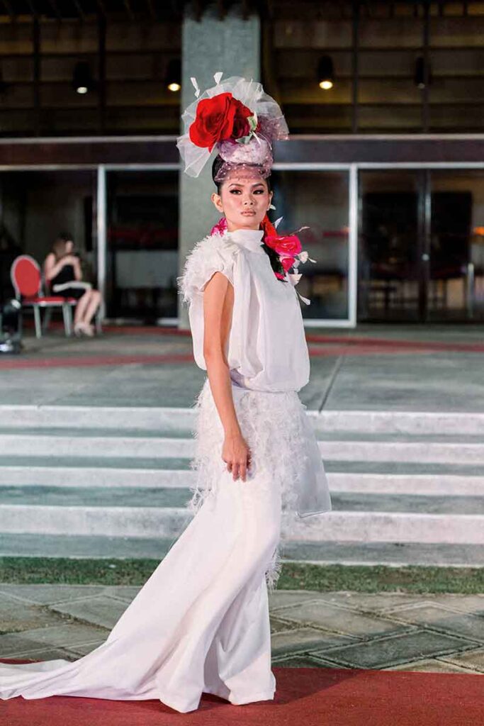 Model in a white Tingting Delfin runway look with a large red flower headpiece